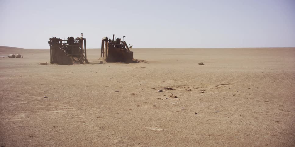 Abandoned, rusted machinery sits decaying in a vast, empty desert as wind blows sand across the desolate landscape