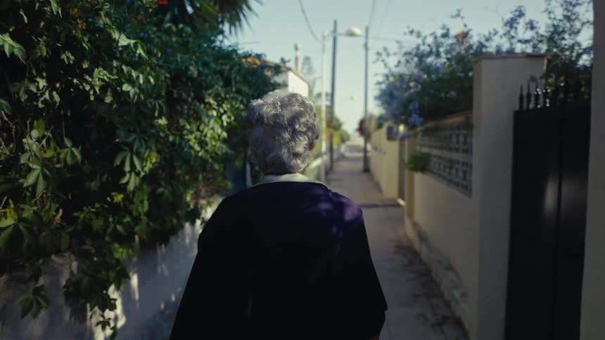 From behind, an elderly woman with grey hair slowly walks down a narrow, sunlit alleyway blooming with flowers