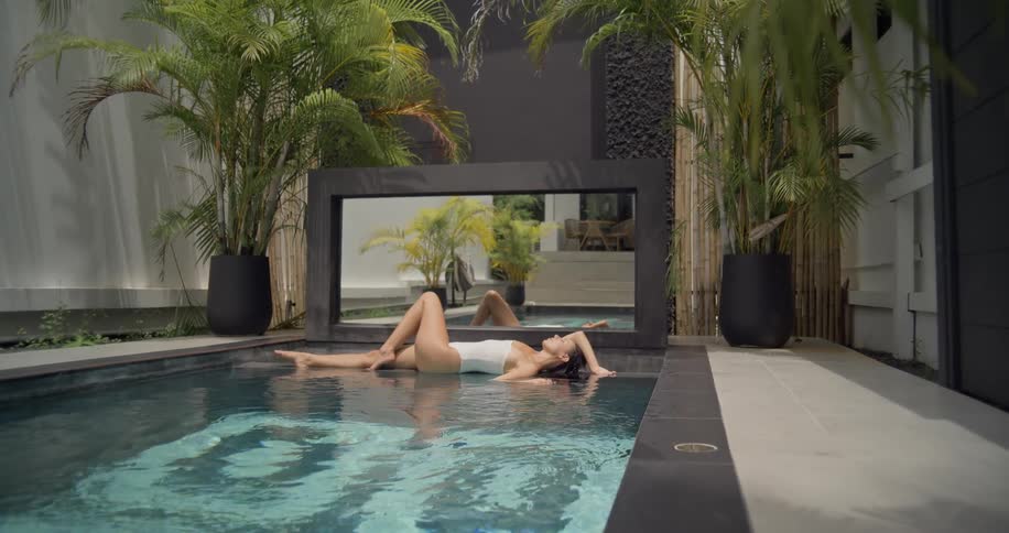 A serene woman in a white one-piece swimsuit relaxes in the tranquil water of a luxurious private pool surrounded by lush tropical plants