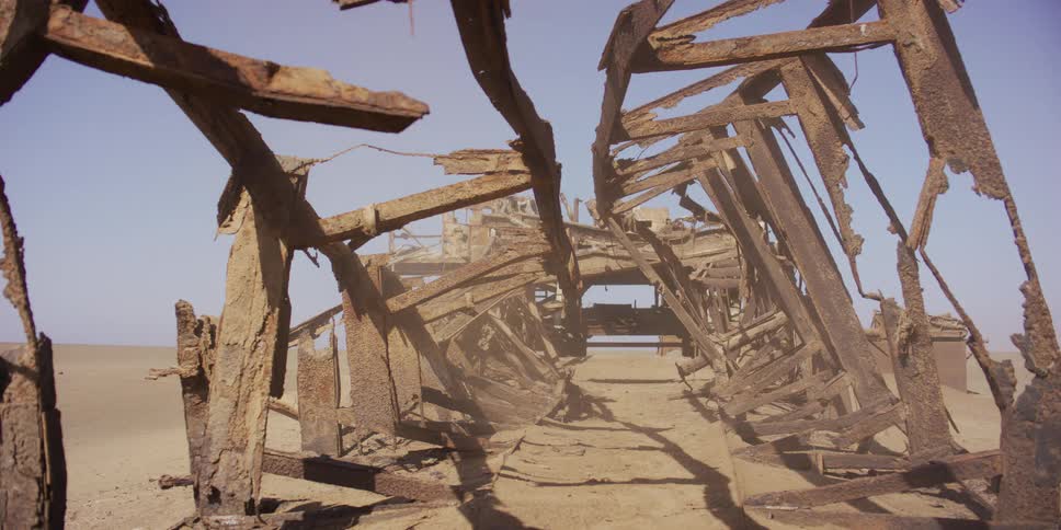 A steady shot from inside the rusted skeleton of an abandoned industrial machine as sand blows in the desert wind