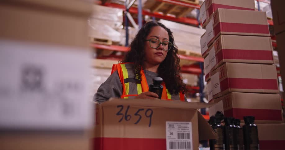A female warehouse worker carefully inspects and packages small bottles into a cardboard box for shipping