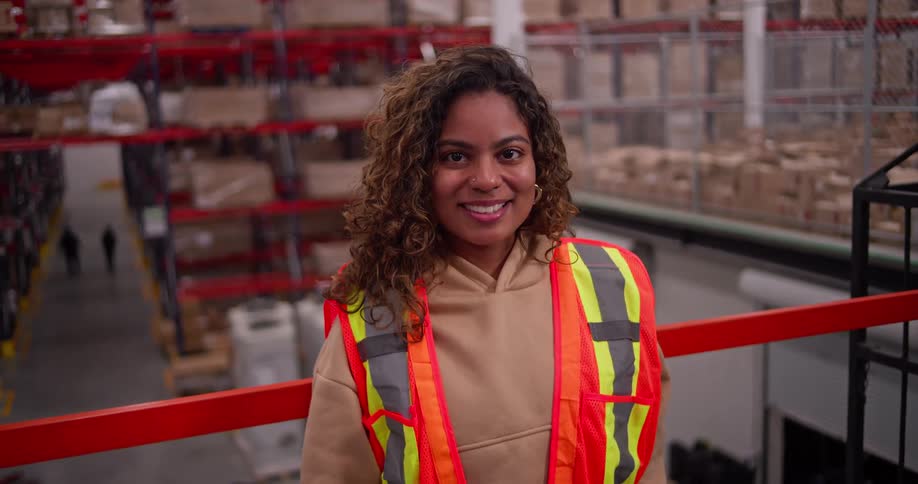 Portrait of a confident young female worker in a safety vest smiling in a large industrial warehouse