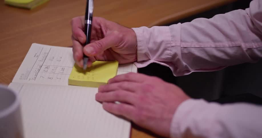 A diligent man in glasses writes important notes on a yellow sticky pad while working at his office desk