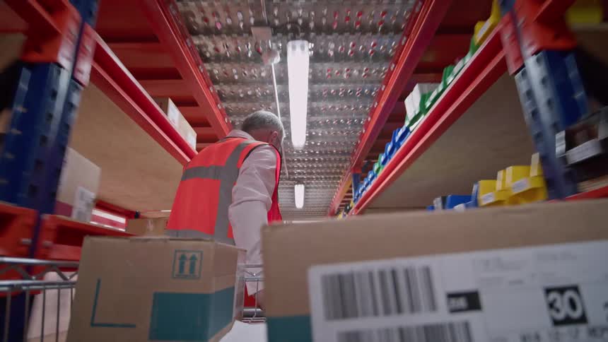 POV shot from a cart as a warehouse worker pushes it down a long aisle between tall shelves of inventory