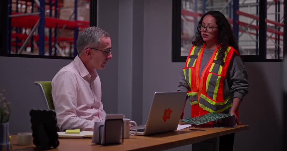 A warehouse manager and female worker discuss logistics in an office overlooking the storage facility