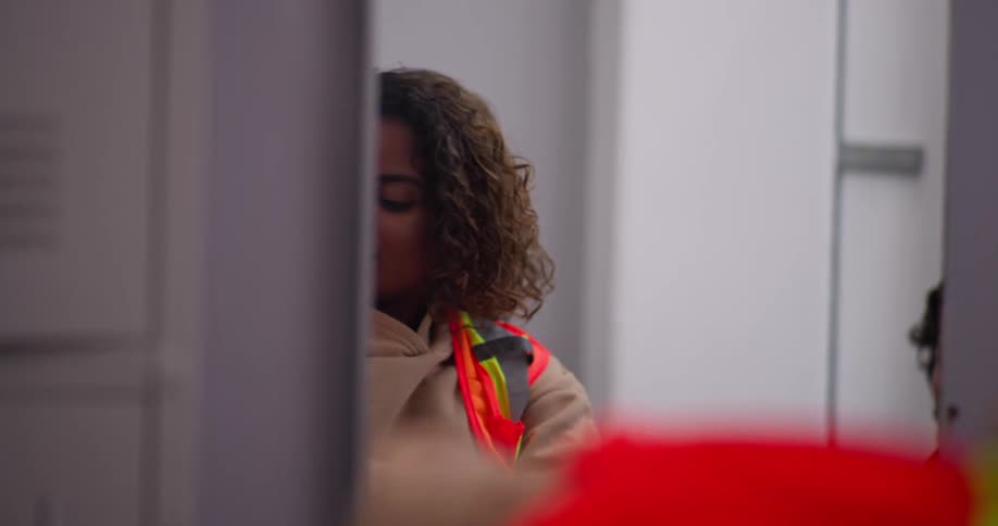A young man chats with a coworker as she puts on her high-visibility vest in the locker room before their shift