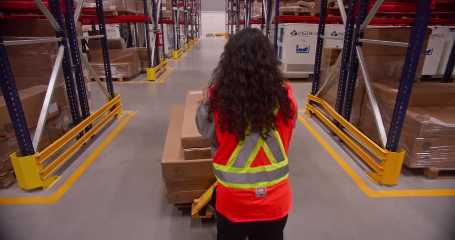 A female worker pushes a pallet jack loaded with boxes down a long aisle in a massive, well-stocked distribution warehouse