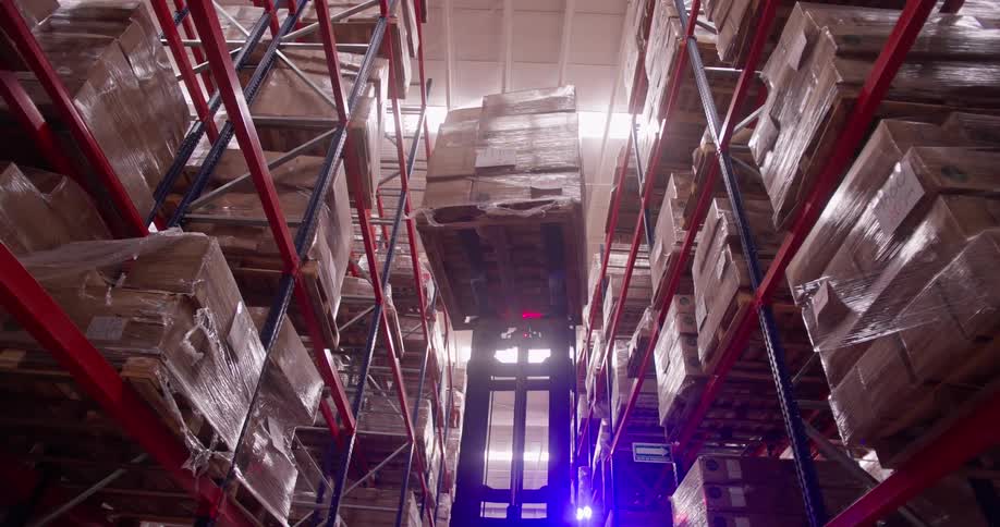 From a low angle, a high-reach forklift retrieves a large pallet of goods from a towering warehouse shelf and lowers it down the aisle