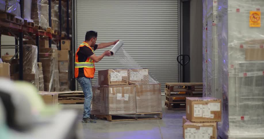 A warehouse worker in a safety vest wraps a pallet of cardboard boxes with stretch film for shipping