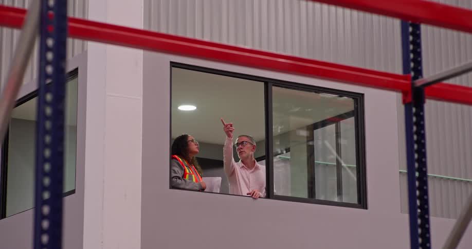 A female warehouse supervisor and a male manager review documents and discuss operations while observing the facility from an elevated office window