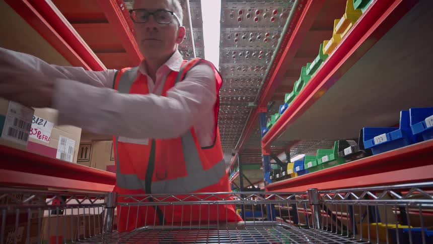 Point-of-view from a trolley as a male warehouse worker stops to check his clipboard, signs for a package, and places it on a shelf