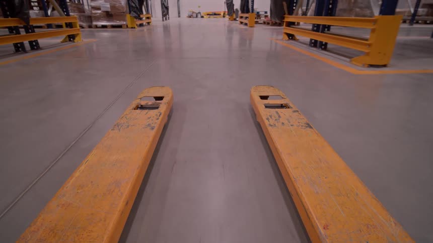 A point-of-view shot from a pallet jack as it lifts a pallet of boxes and moves backward through a large warehouse