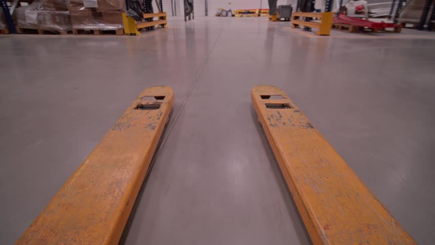 First-person view of a pallet jack navigating a warehouse aisle and preparing to lift a pallet of merchandise