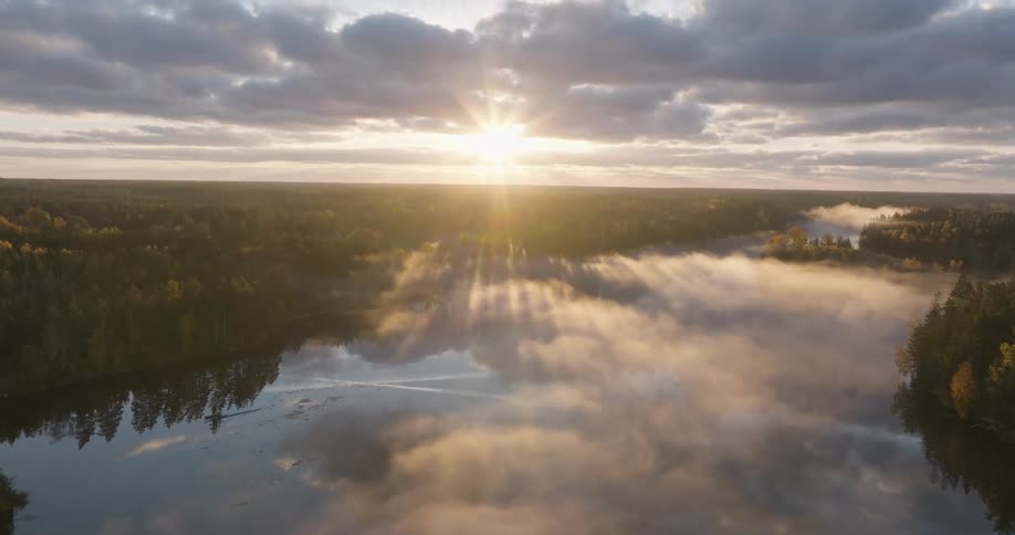 Breathtaking aerial view of a misty lake at sunrise with golden sun rays piercing through the fog over an autumn forest