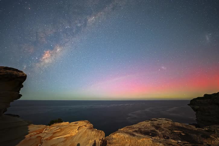 A stunning time-lapse captures the Milky Way and the colorful Aurora Australis dancing in the night sky over the ocean from a rocky coastal viewpoint