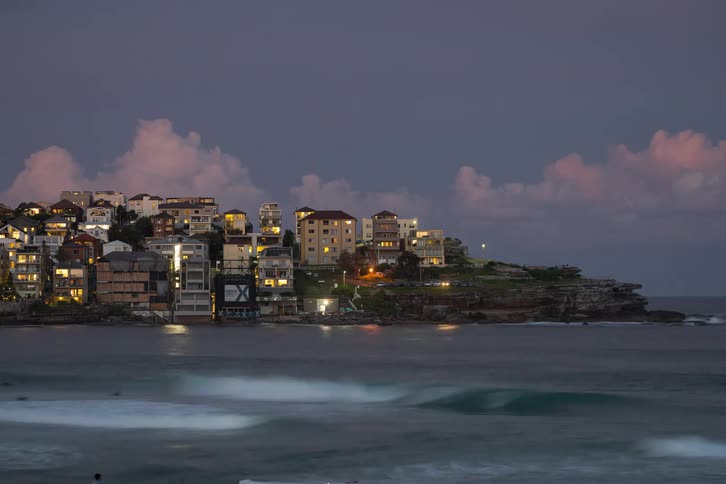 Time-lapse of misty waves rolling ashore at Bondi Beach as the coastal town lights up at dusk under a beautiful cloudy sky