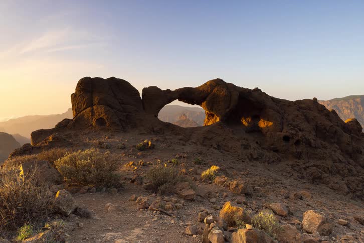A time-lapse captures the sunrise casting warm golden light and long shadows across the La Ventana del Nublo rock arch in Gran Canaria