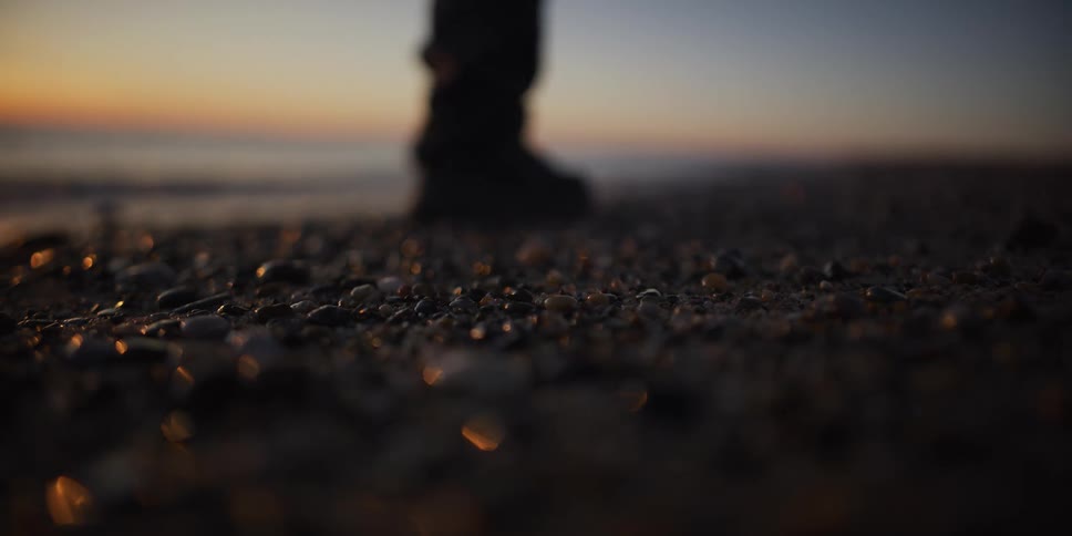 A person in boots slowly walks across a pebble beach and stops to watch the serene sunset over the calm ocean