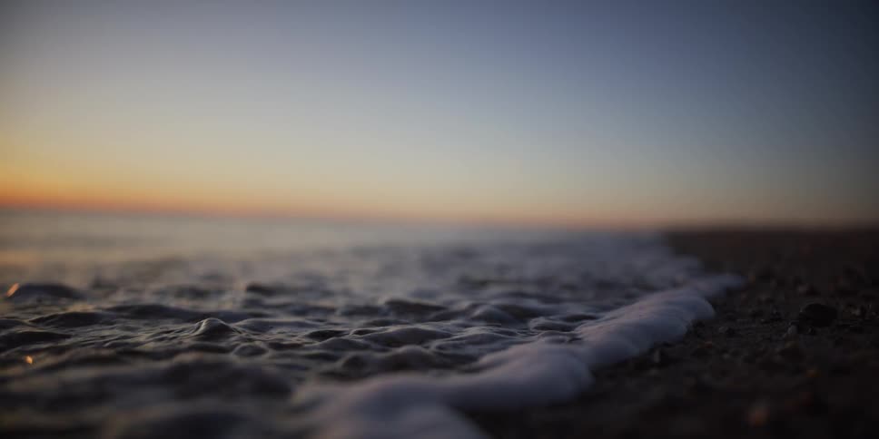 A serene, slow-motion, close-up of a gentle wave washing over a pebbly beach during a beautiful sunset