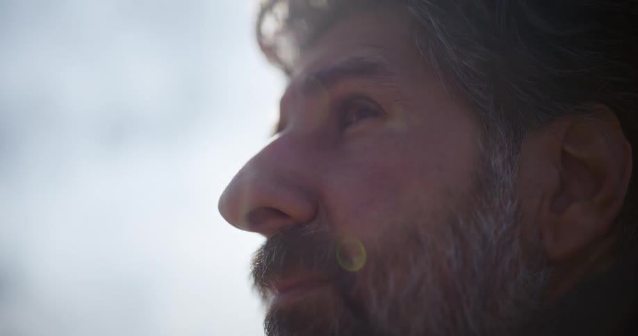A contemplative middle-aged man with a gray beard looks up at the sky in a sun-drenched close-up shot