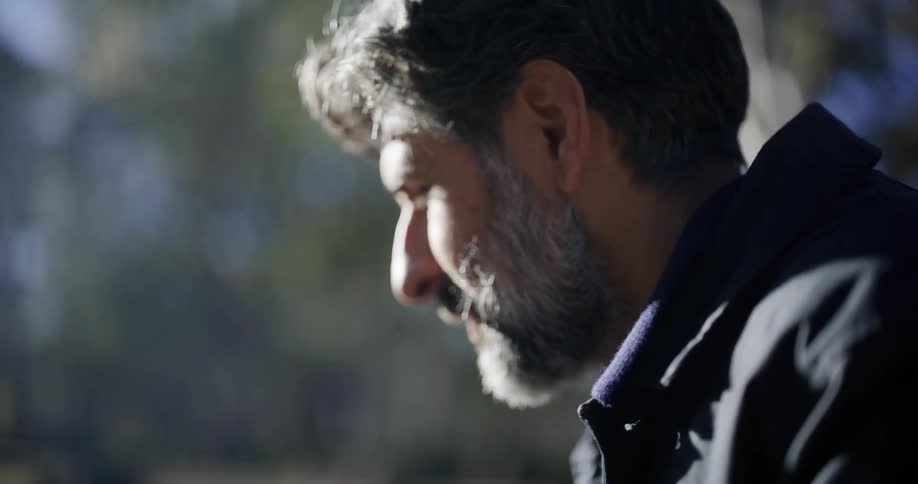 Slow motion close-up of a rugged man with a grey beard drinking a steaming hot beverage from a mug in the morning sun