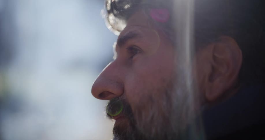 A pensive middle-aged man with a salt-and-pepper beard is seen in a sun-drenched, out-of-focus close-up profile shot