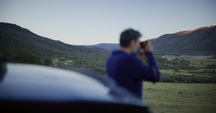 A man on a scenic road trip stops to use binoculars and admire the vast mountain valley during the golden hour