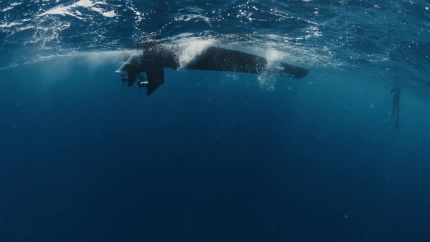 Sunlight filters through the choppy ocean surface, revealing the silhouette of a boat and a freediver from an underwater perspective