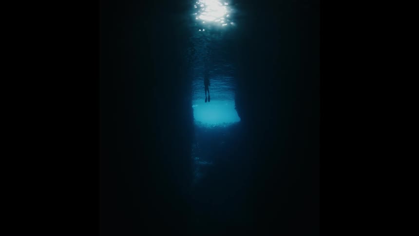 A silhouetted freediver ascends through a dark underwater canyon towards the light shimmering on the ocean's surface