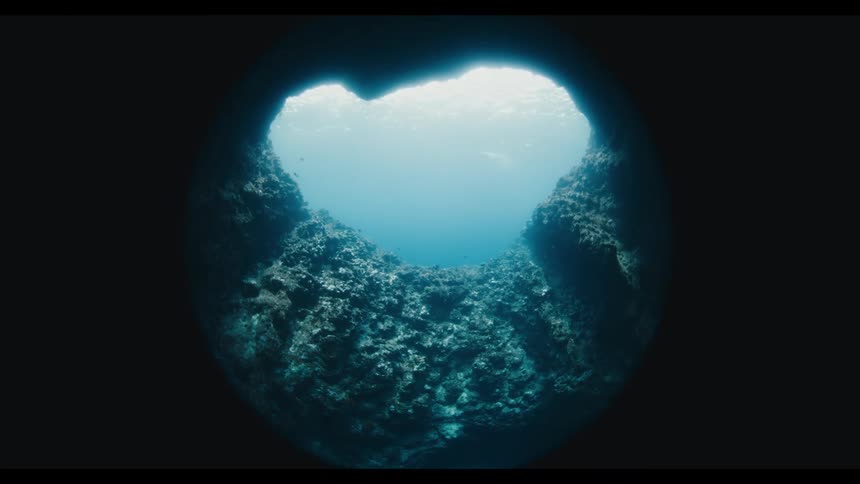 A fisheye view from the dark depths of an underwater cave looking up at the bright, shimmering ocean surface