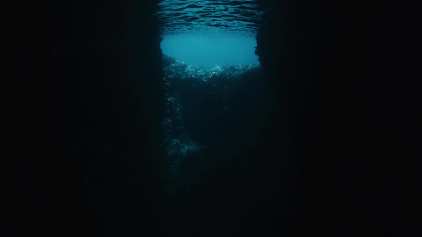 An eerie underwater perspective from inside a dark sea cave looks up towards the bright, rippling surface of the ocean