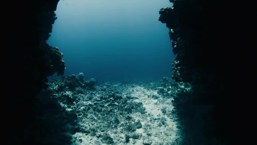 Slowly drifting out of a dark underwater sea cave to reveal the beautiful coral reef and deep blue ocean