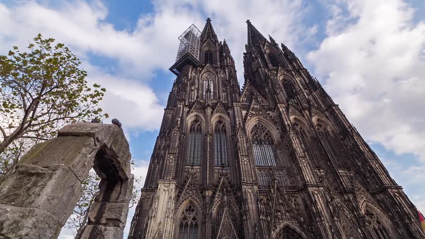 A dramatic low-angle timelapse shows clouds rolling over the magnificent Gothic facade of the Cologne Cathedral in Germany, with ancient ruins in the foreground