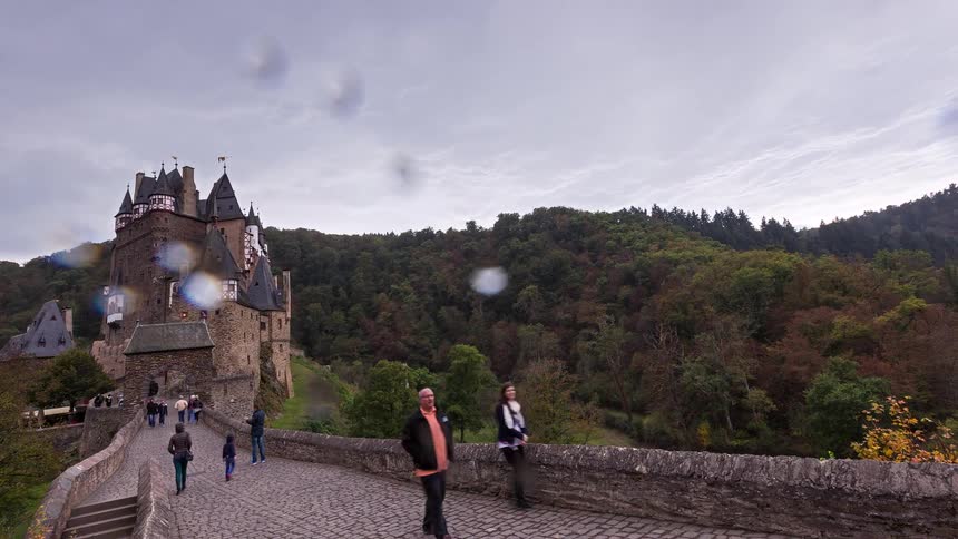 A time-lapse of tourists walking across the cobblestone bridge to the historic Eltz Castle in Germany, surrounded by a lush autumn forest under a cloudy sky