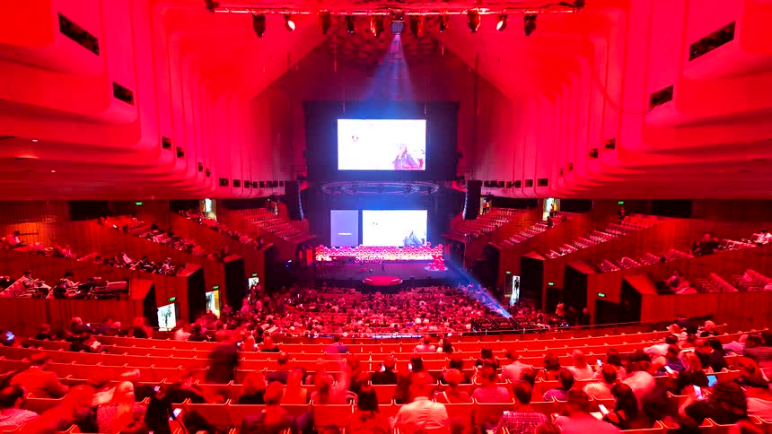 Time-lapse of an audience filling a grand auditorium, bathed in vibrant red light, for a large conference or presentation