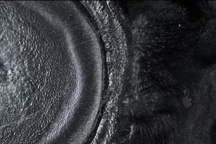 A viscous, sparkling black and silver fluid moves slowly, creating waves and textured shapes in an abstract macro shot