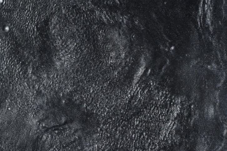 Molten silver-black paint with glitter particles slowly moves and solidifies into a dark, textured background