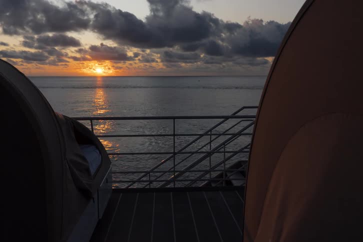 A serene time-lapse captures the sun rising over a calm ocean, illuminating the cloudy sky as seen from the deck of a boat