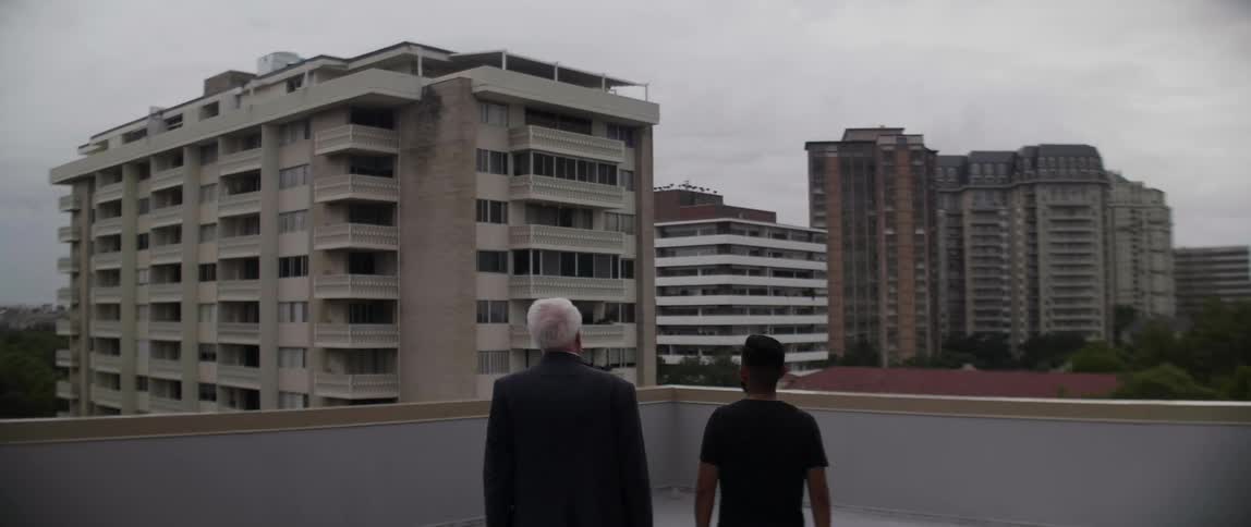 Two men stand on a balcony looking at buildings
