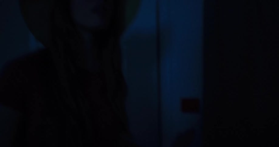 A young woman wearing a hat cautiously enters a dark room and turns on the lights, revealing a hotel room interior