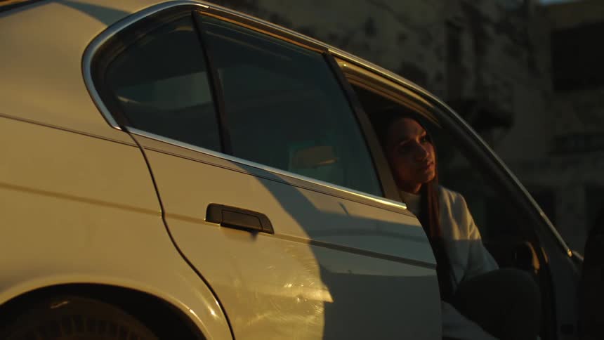 A thoughtful woman sits in the driver's seat of her car with the door open, illuminated by the warm glow of the golden hour sun