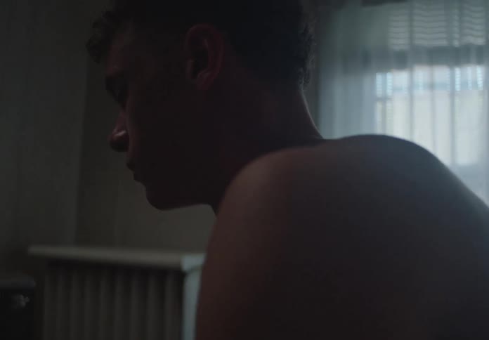 Slow-motion close-up of a shirtless young man sitting alone in a dimly lit room with a pensive expression