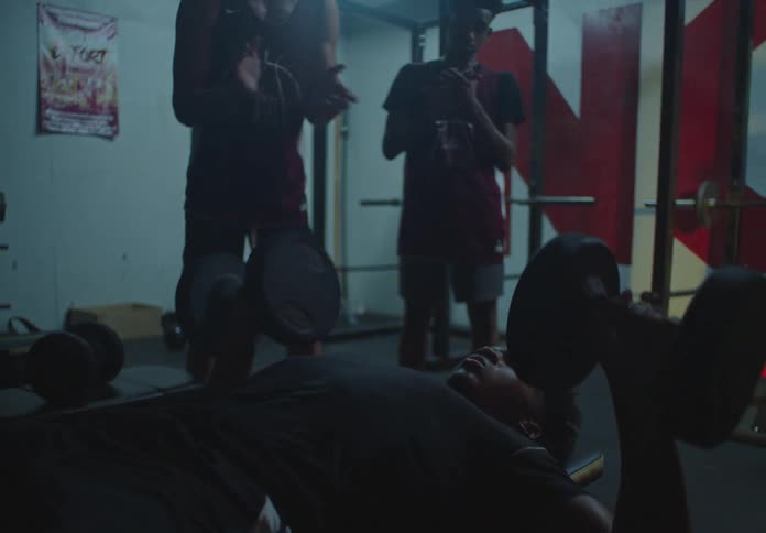 A young basketball player lifts heavy dumbbells on a bench press in while his teammates watch and encourage him in a gritty gym