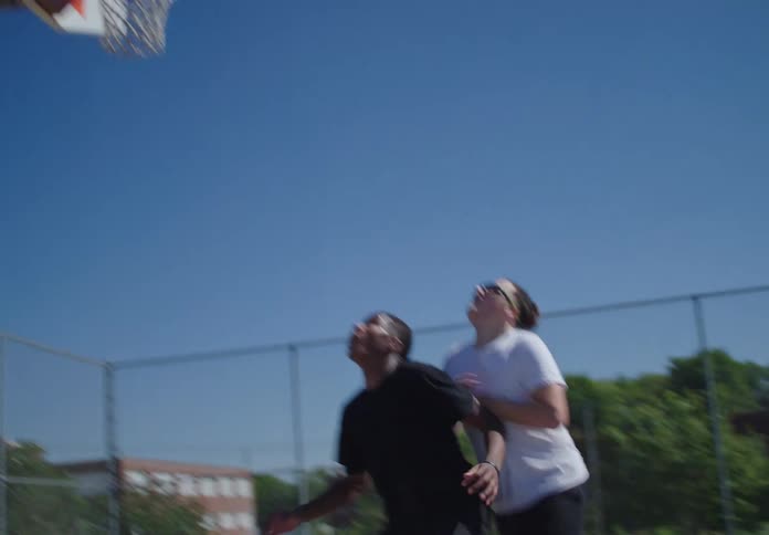 A young man makes a jump shot over a defender and scores in on a sunny outdoor basketball court