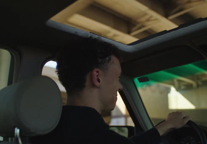 A young man with curly hair drives his car under a highway overpass as sunlight flickers through the sunroof