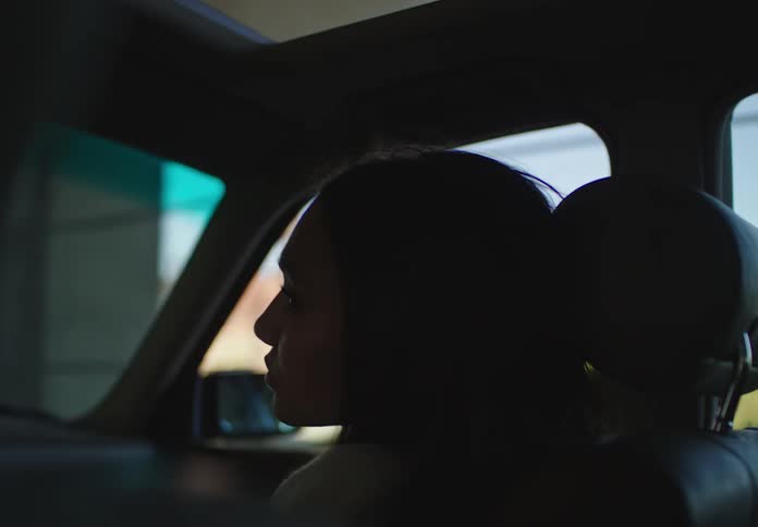A contemplative woman in silhouette looks out the window from the passenger seat of a moving car as the scenery passes by