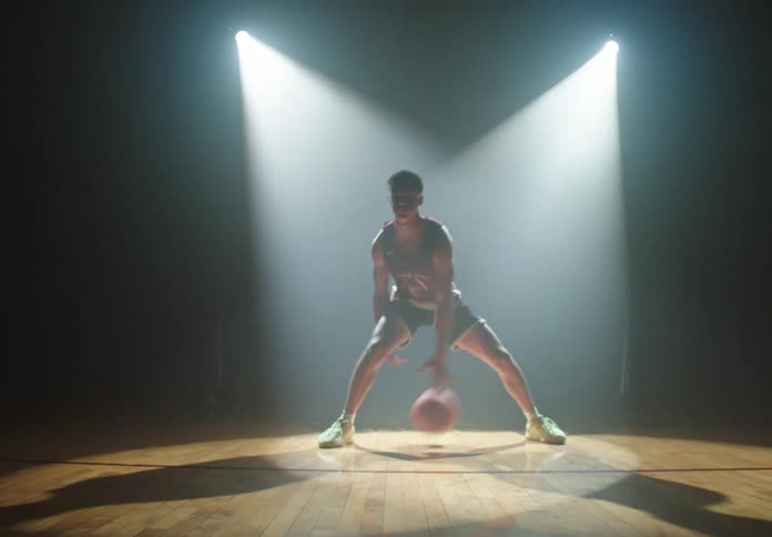 Dolly in on a lone basketball player dribbling with skill under two dramatic spotlights on a dark, reflective court