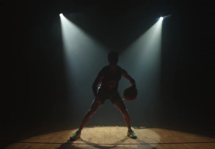 A focused basketball player dribbles a ball on a court under two dramatic spotlights in a dark, smoky arena