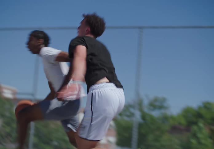 A young man dribbles past a defender and impressively dunks the basketball in slow motion on a sunny outdoor court