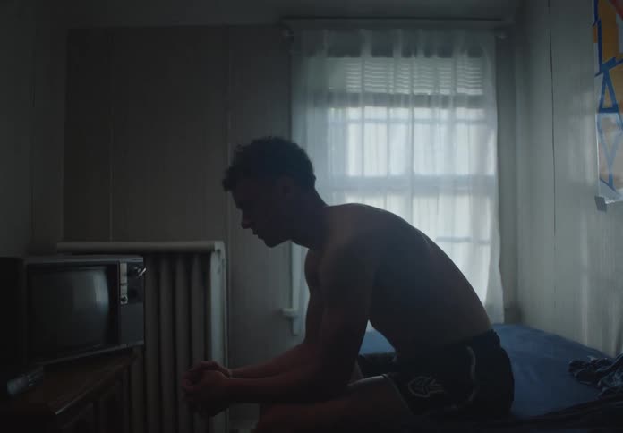 A pensive young man sits shirtless on the edge of his bed in a dimly lit room, lost in his thoughts